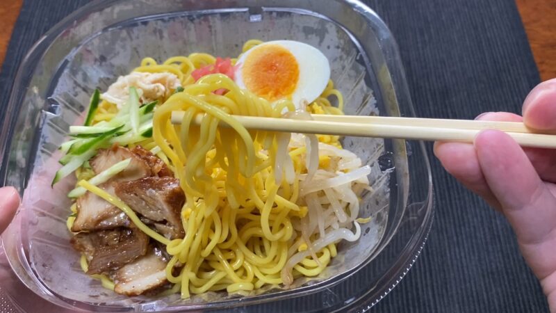 ローソン冷やし中華を箸で持ち上げた様子。チャーシューやきゅうり、錦糸卵、半熟卵の具材が見える