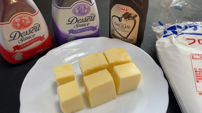 白い皿にカットされたリッチチーズケーキが6個並び、背後にはストロベリー、ブルーベリー、チョコレートのデザートソースとホイップクリームのパッケージが置かれている。デザートのアレンジを楽しむ準備が整っている様子。