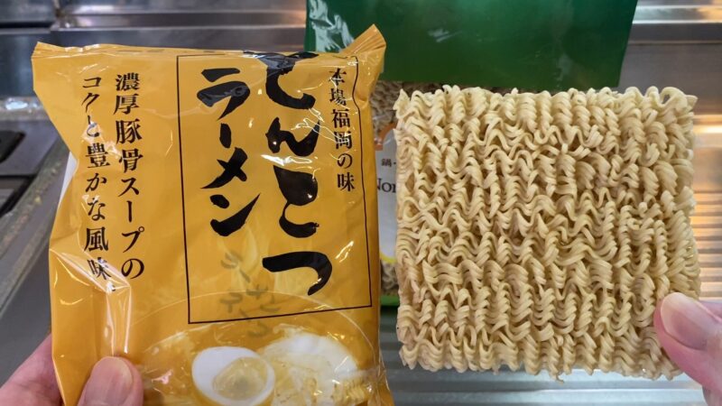 黄色いパッケージのとんこつラーメンと、中身の乾燥麺を並べて持っている様子。麺は波打つ形状で、パッケージには「濃厚豚骨スープのコクと豊かな風味」と記載されている。