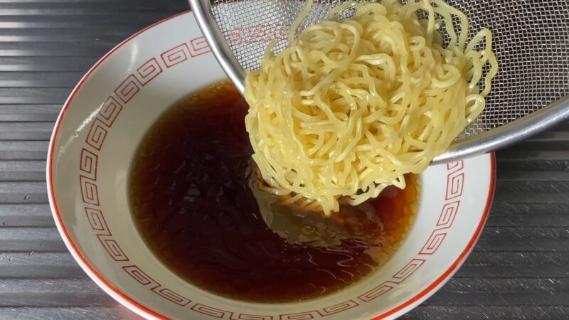 醤油スープの入った丼に、湯切りしたウェーブ麺を入れている瞬間。麺が艶やかに光っている。