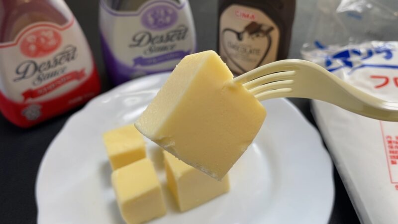 フォークに刺さった一口サイズのチーズケーキのアップ。後ろにデザートソースのボトルがぼんやり写る。