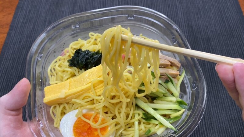 セブンイレブンの冷やし中華を箸で持ち上げ、麺のつややかな質感を映した実食の瞬間。