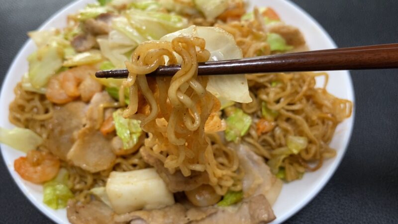箸で持ち上げた業務スーパーのマレーシア産ノンフライ乾麺を使ったシーフード焼きそば。麺にキャベツとソースが絡み、背景にはエビや豚肉が見える。