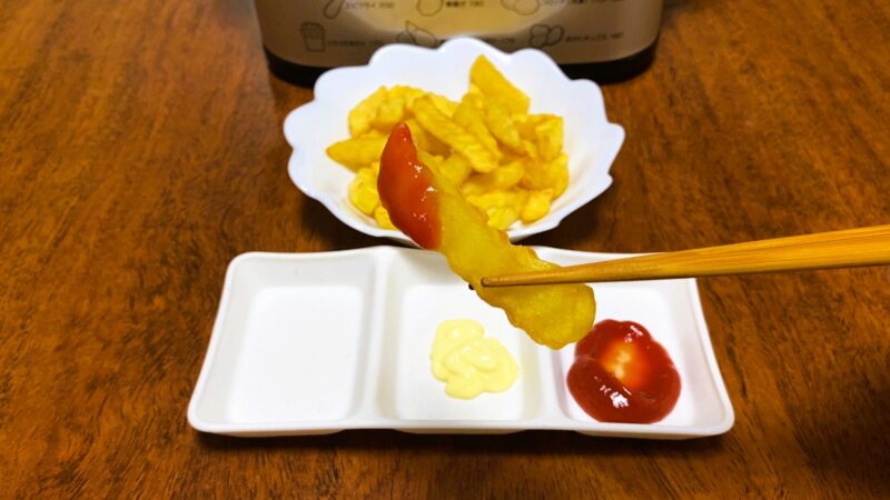 ケチャップとマヨネーズが添えられた白い小皿に、ケチャップをつけたフライドポテトを箸で持ち上げているシーン。