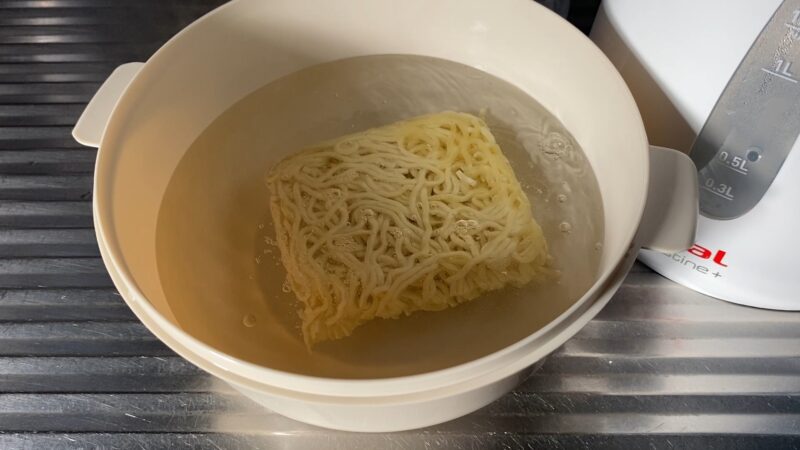 耐熱容器に入れた冷凍ウェーブ麺を熱湯でほぐしている様子。麺が徐々にほぐれ始めている。