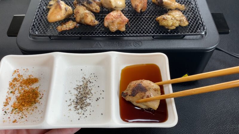 電気七輪で焼いた焼き鳥をタレにつけているところ。調味料皿には七味・胡椒・タレが用意されている。