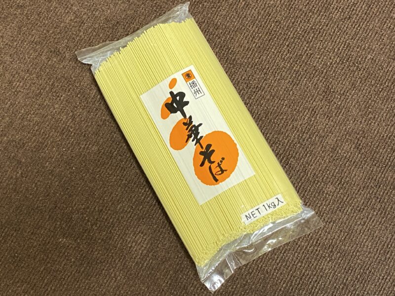 透明袋に入った「播州 中華そば」1kgパッケージ。黄色い細麺がぎっしり詰まっており、中央には白地にオレンジの丸と黒文字のラベルが貼られている。