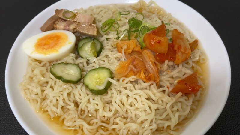 白い器に盛られた冷やし麺。醤油ベースの冷たいスープに、チャーシュー、半熟卵、キムチ、きゅうり漬け、刻みネギがトッピングされている。ノンフライの縮れ平麺が涼しげな印象を与える一品。