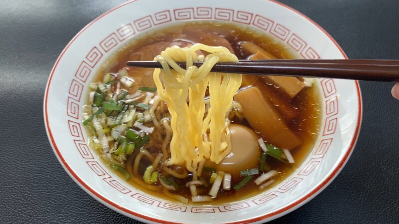 醤油スープのラーメンを箸で持ち上げている様子（具材は味玉、メンマ、刻みネギ）