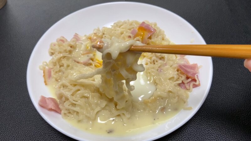 カルボナーラソースと半熟卵が絡んだノンフライ乾麺を箸で持ち上げたところ。ベーコンも見える。