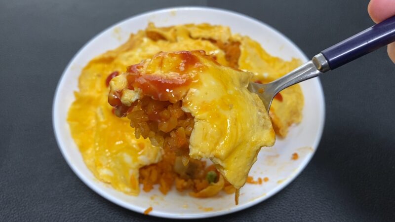 ケチャップをかけたチキンライス入りオムライスをスプーンですくったアップ写真。とろとろの卵とチキンライスが層になっている。