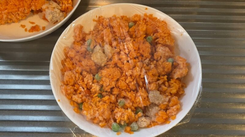 白い器に冷凍チキンライスを入れてラップをかけた状態。中にはグリーンピースや鶏肉のかたまりが見える。