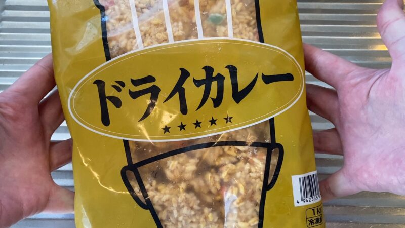 業務スーパーの冷凍ドライカレー1kgパッケージを両手で持っている様子。黄色いパッケージでシンプルなデザイン。