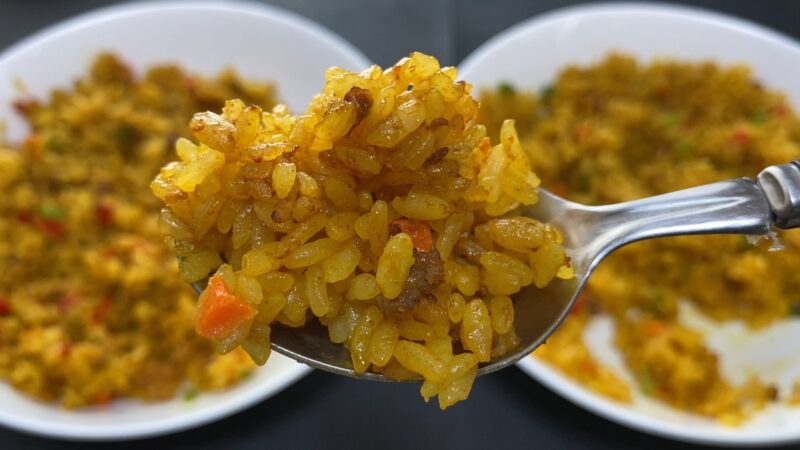 フライパンで炒めたドライカレーをスプーンですくった接写。背景には白い皿に盛られたドライカレーが2皿見える