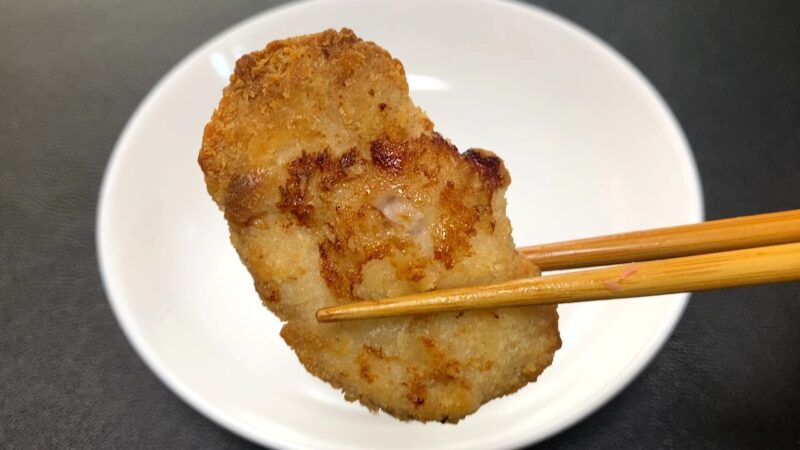 ノンフライヤーで加熱した業務スーパー「肉厚 極旨とんかつ」を箸で持ち上げた写真。衣はやや硬く、油で揚げたときのような香ばしさは少ないが、軽い仕上がり。