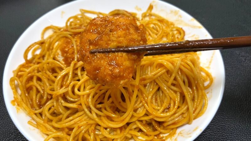 ミートソースを絡めたパスタの上で、チキンカツを箸で持ち上げた瞬間。ソースが衣にしっかり絡み、食欲をそそる見た目。