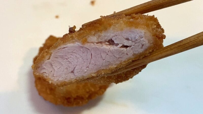 箸で持ち上げた業務スーパー「肉厚 極旨とんかつ」の断面。衣の中にやわらかい豚肉が見え、肉質のきめ細かさがわかる。