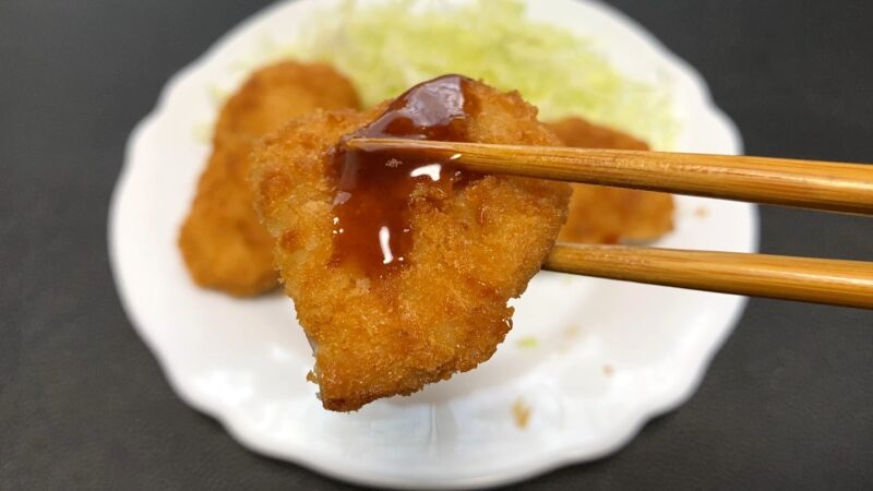 業務スーパー「肉厚 極旨とんかつ」にトンカツソースをかけ、箸で持ち上げた写真。衣のサクサク感と照りのあるソースが食欲をそそる。