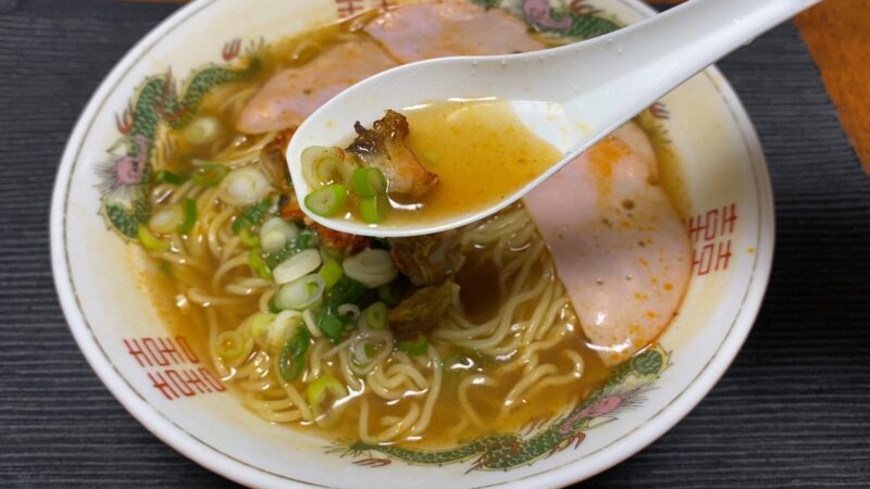 ザリガニ出汁を使ったラーメンのスープ。レンゲに具材をのせたアップで、ほんのり赤みのある澄んだ醤油スープが印象的。