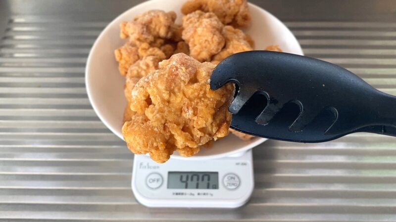 業務スーパー「和風鶏もも唐揚げ」の1個をトングで持ち上げ、スケール上で重さを測っている場面。477gの表示が確認できる。サイズ感の比較用途に適した画像