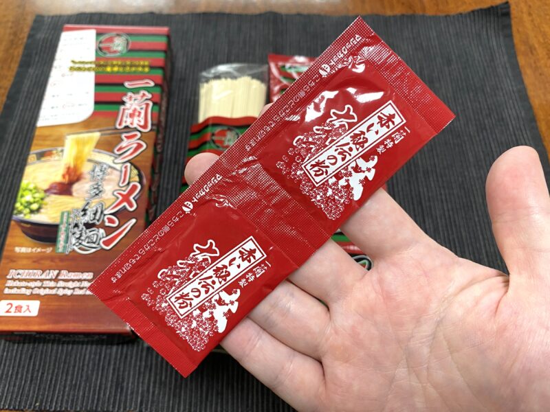 一蘭の赤い秘伝のたれを手に持った写真。辛味調味料の小袋が2つ並んでいる。