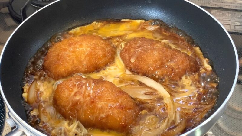 フライパンで業務スーパーの冷凍とんかつを卵と玉ねぎと一緒に煮込んでいる様子。すき焼きのタレを薄めた簡易ダレで、衣にタレが染み込み始めている。