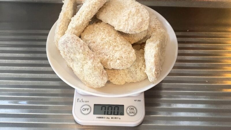 業務スーパー「肉厚 極旨とんかつ」を皿に盛り、キッチンスケールで計量している写真。表示は700グラムで、14枚ほどの冷凍とんかつが積み重なっている。