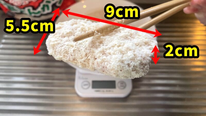 業務スーパー「肉厚 極旨とんかつ」のサイズを計測している写真。縦5.5cm、横9cm、厚み2cmと記載があり、箸で持ち上げた冷凍とんかつの大きさがわかる。