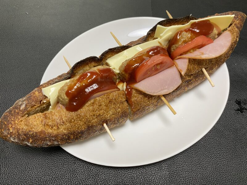 焼上ミニハンバーグをバゲットにサンドしたアレンジ料理。チーズとハム、トマトを重ねたボリューム満点の一品。