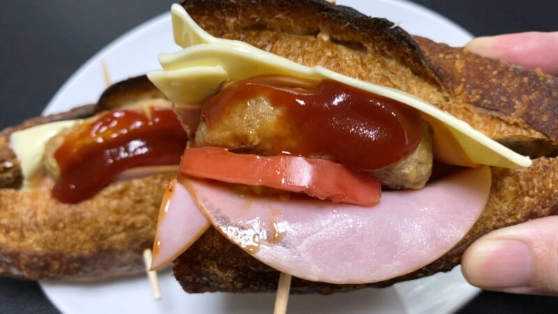 ハム、トマト、ミニハンバーグ、チーズをはさんだバゲットサンドを正面から撮影。全体のボリューム感がわかる