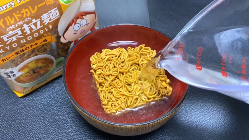 東京拉麺に熱湯を注いでいる調理シーン。メジャーカップからお湯を注ぎ、麺がふやけていく様子が写っている。