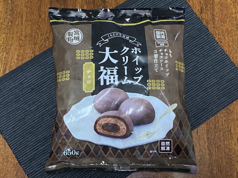 業務スーパー「ホイップクリーム大福(チョコ)」のパッケージ。黒いランチョンマットの上に置かれており、自然解凍・3層仕立ての説明が見える。