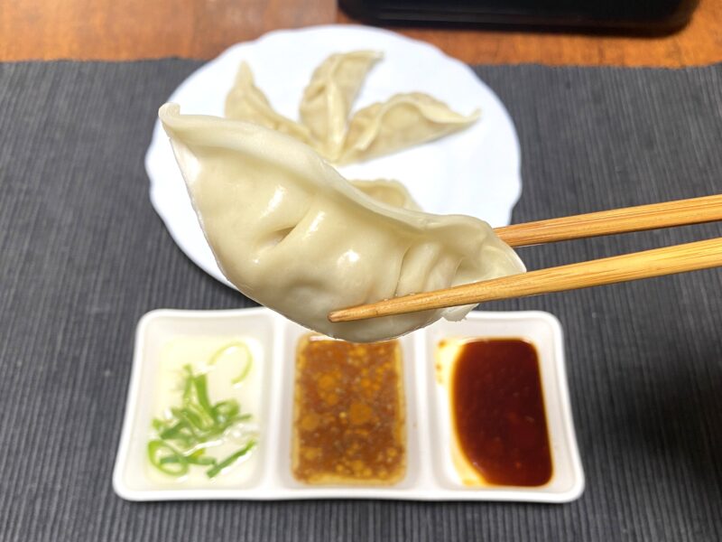 箸で持ち上げた蒸した豚肉餃子のアップ写真で、なめらかで光沢のある皮の質感が見える。奥には3種類のタレが配置されている。