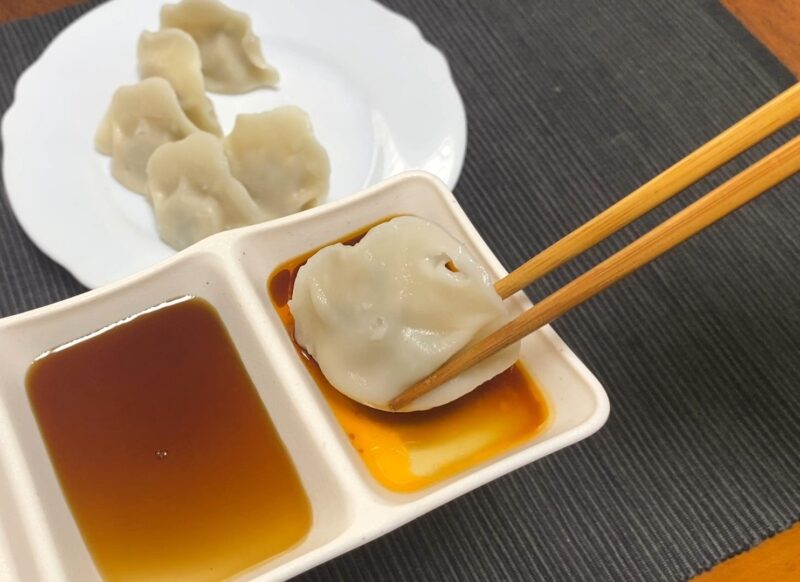醤油とラー油のタレにつけている茹でた三鮮水餃子。タレ皿と残りの餃子が背景に写っている。