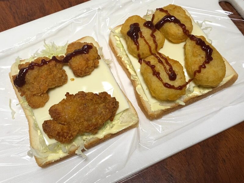 食パンにキャベツ、スライスチーズ、マヨネーズをのせ、その上に唐揚げとナゲットを並べたサンドイッチの準備段階の写真。