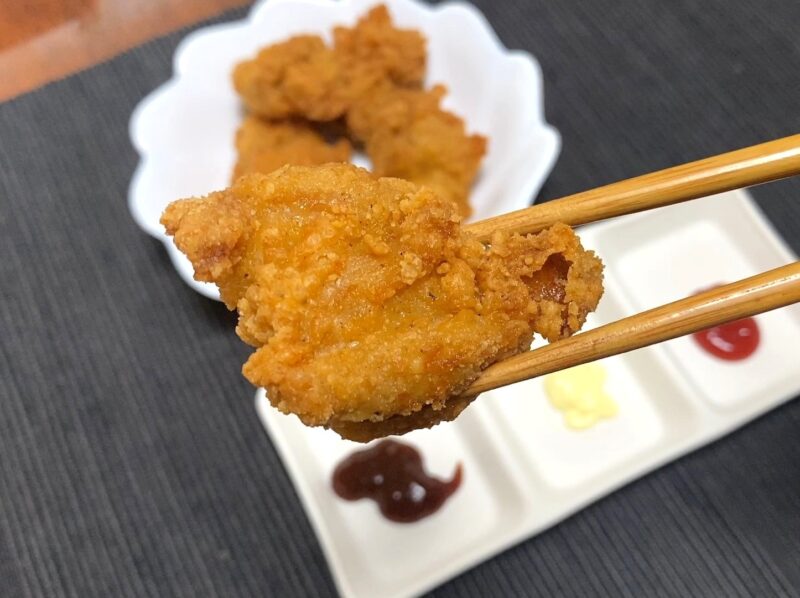 油で揚げた業務スーパー「お肉にこだわった美味しい唐揚げ（タイ産）」を箸で持った試食シーン。後ろにソース皿が写っている。