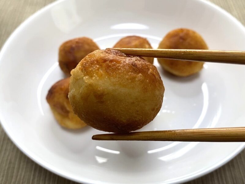 箸で持ち上げた、こんがり焼けた冷凍たこ焼きのクローズアップ写真。外側はカリッと焼き目が付き、中はもっちり食感のたこ焼きが皿の上に並んでいる様子。