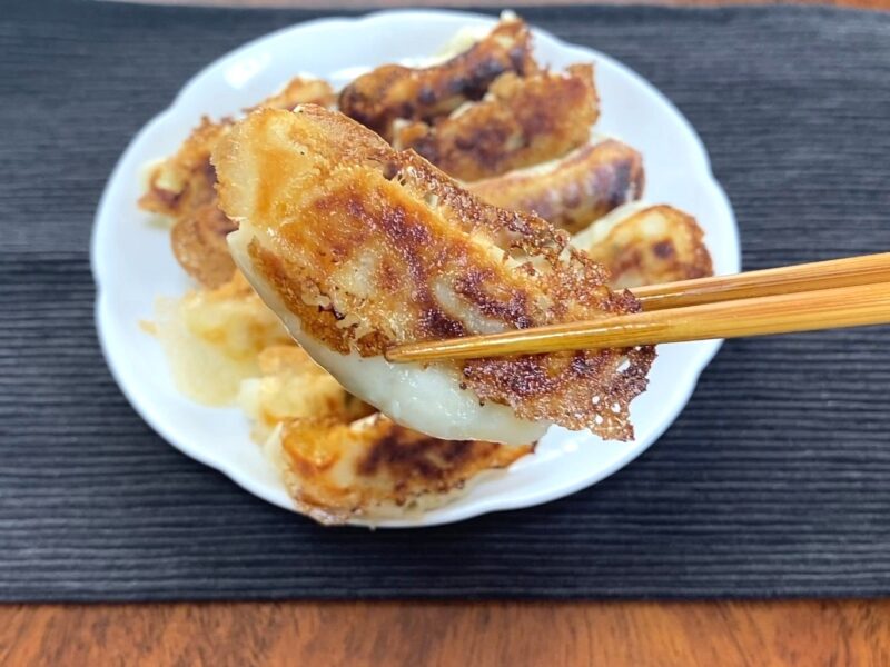 焼き上がったコスモス羽根つきギョーザを箸で1個つまんだところ。こんがりした焼き色とパリパリの羽根